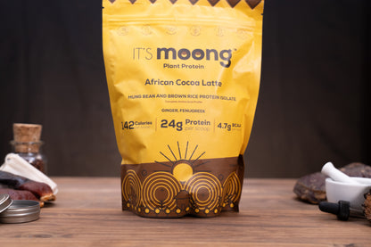 A yellow stand-up pouch of It's Moong Plant Protein African Cocoa Latte stands on a wooden surface against a dark background, with small ingredients and tools placed around it.