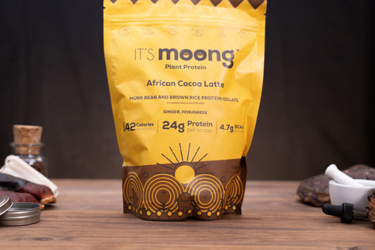 A yellow stand-up pouch of It's Moong Plant Protein African Cocoa Latte stands on a wooden surface against a dark background, with small ingredients and tools placed around it.