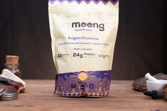 A stand-up pouch of It's Moong Plant Protein in Belgian Chocolate flavor, standing on a wooden surface with a dark, rustic background and small props like a glass bottle, metal lid, and mortar and pestle.