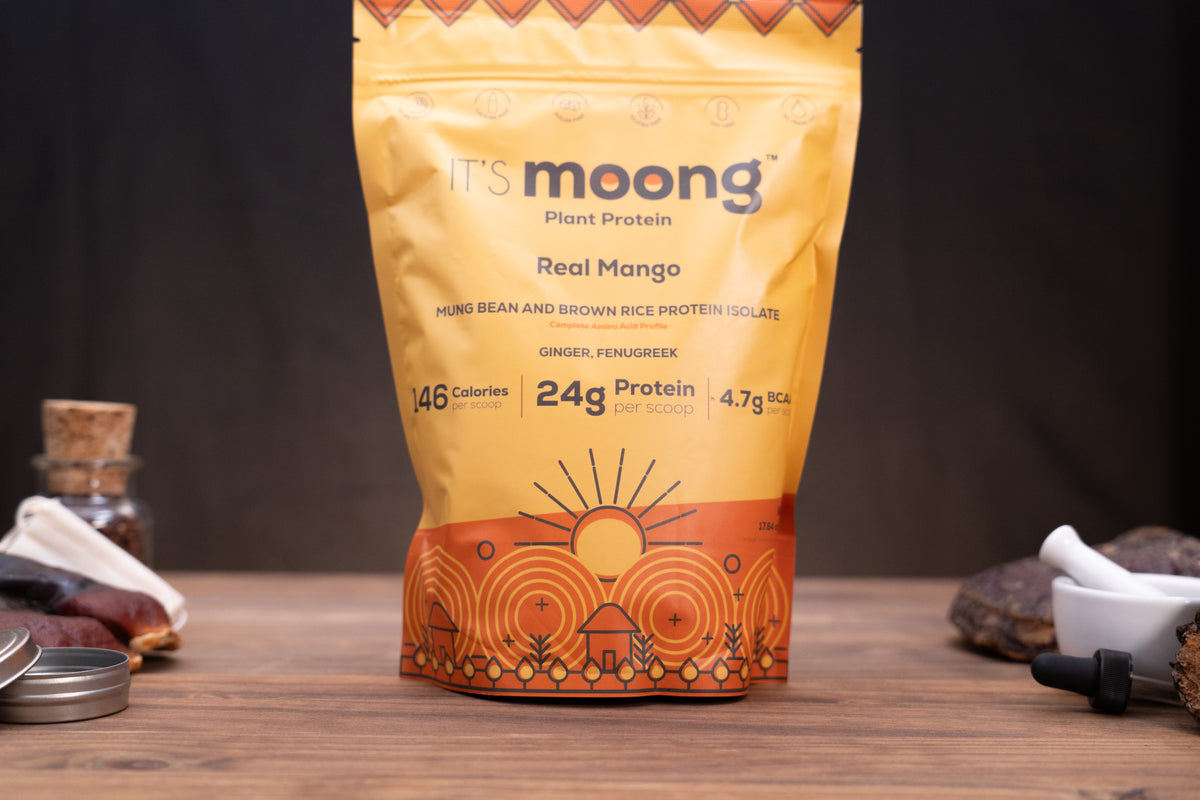 A stand-up pouch of It's Moong Real Mango plant protein powder, featuring a warm yellow and orange design with a sun illustration, displayed on a wooden surface with rustic ingredients in the background.
