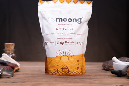 A stand-up pouch of It's Moong Unflavored Plant Protein powder on a wooden tabletop, with blurred jars and ingredients around it against a dark background.