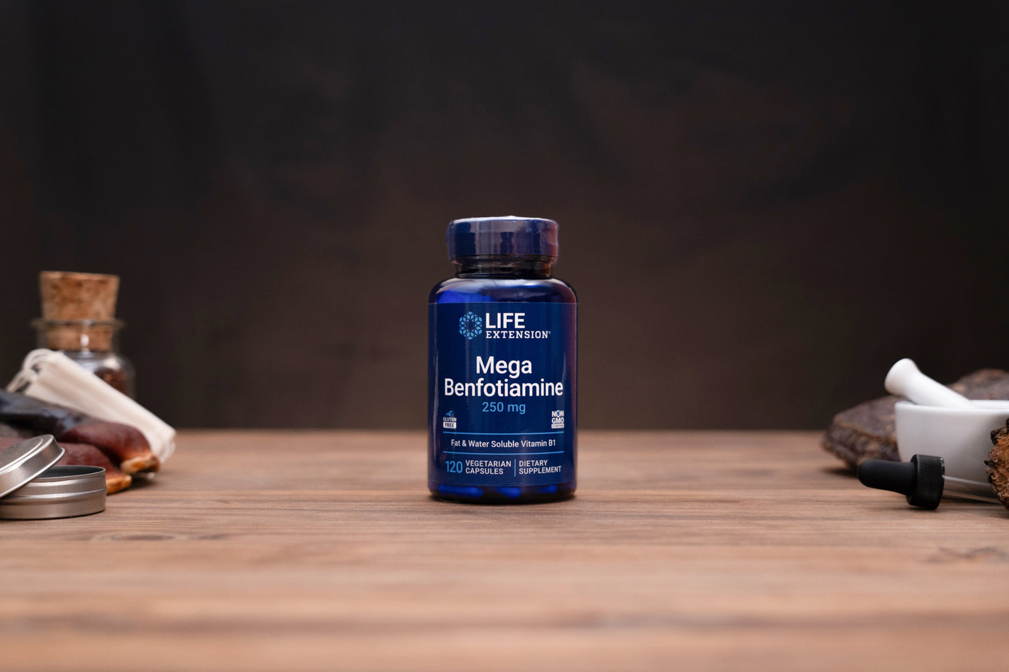 A bottle of Life is Now Mega Benfotiamine 250 mg dietary supplement, set on a wooden table with a dark, moody background and small herbal props on either side.