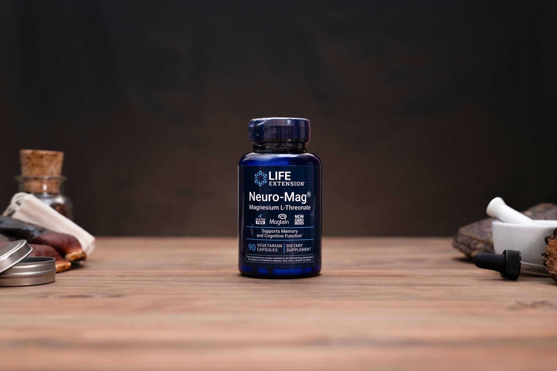 A dark blue bottle of Life Extension Neuro-Mag Magnesium L-Threonate supplement, centered on a wooden tabletop with blurred herbal and laboratory props in the background.