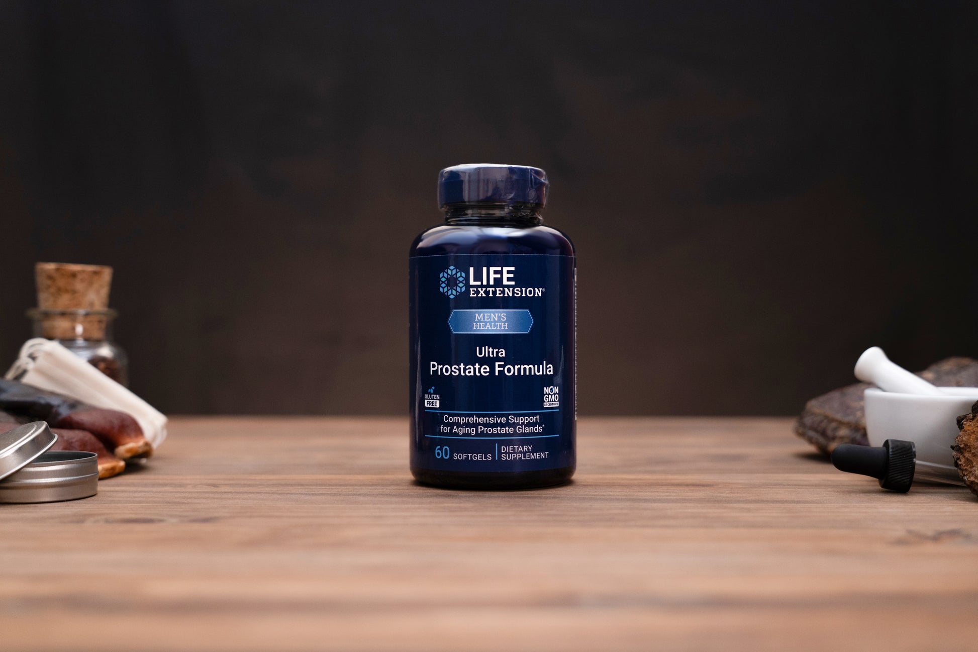 A dark blue bottle of Life Extension Ultra Prostate Formula stands centered on a wooden tabletop, with blurred herbal and laboratory props in the background and foreground.