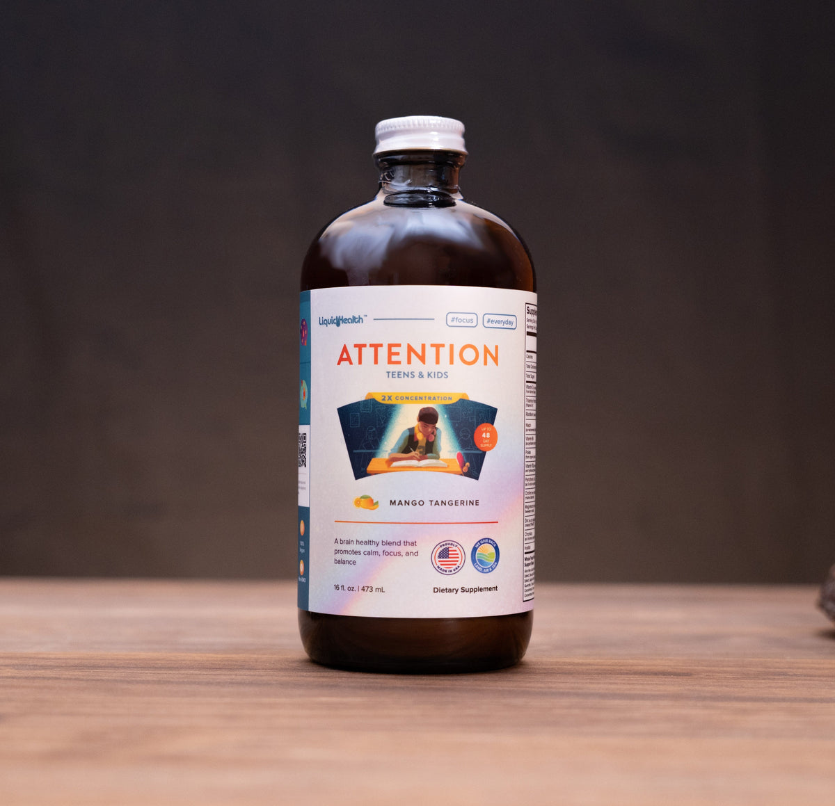 An amber glass bottle of Attention + Focus supplement with a white cap and a white label, standing on a wooden surface with blurred natural props in the background.