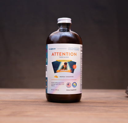 An amber glass bottle of Attention + Focus supplement with a white cap and a white label, standing on a wooden surface with blurred natural props in the background.