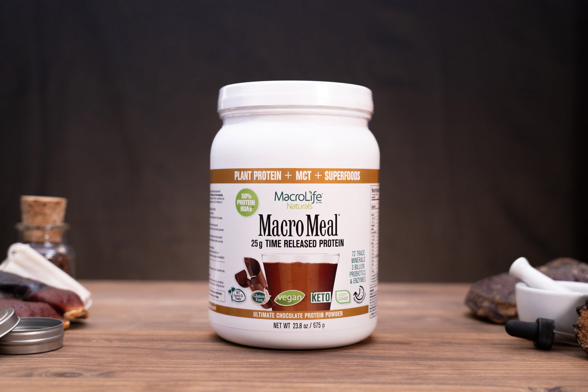 A white plastic tub of MacroLife MacroMeal plant-based protein powder sits on a wooden tabletop against a dark backdrop. The label indicates chocolate flavor and includes a white lid, with small surrounding props like a metal tin lid, a small bottle, and a dropper on both sides.