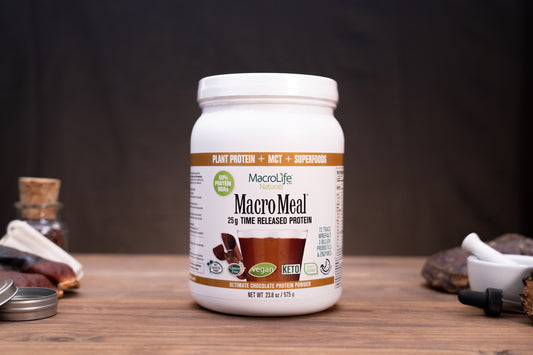 A white plastic tub of MacroLife MacroMeal plant-based protein powder sits on a wooden tabletop against a dark backdrop. The label indicates chocolate flavor and includes a white lid, with small surrounding props like a metal tin lid, a small bottle, and a dropper on both sides.