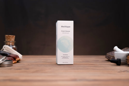 Mad Hippie Cream Cleanser product box standing upright on a wooden surface against a dark background, with small styled props on both sides.
