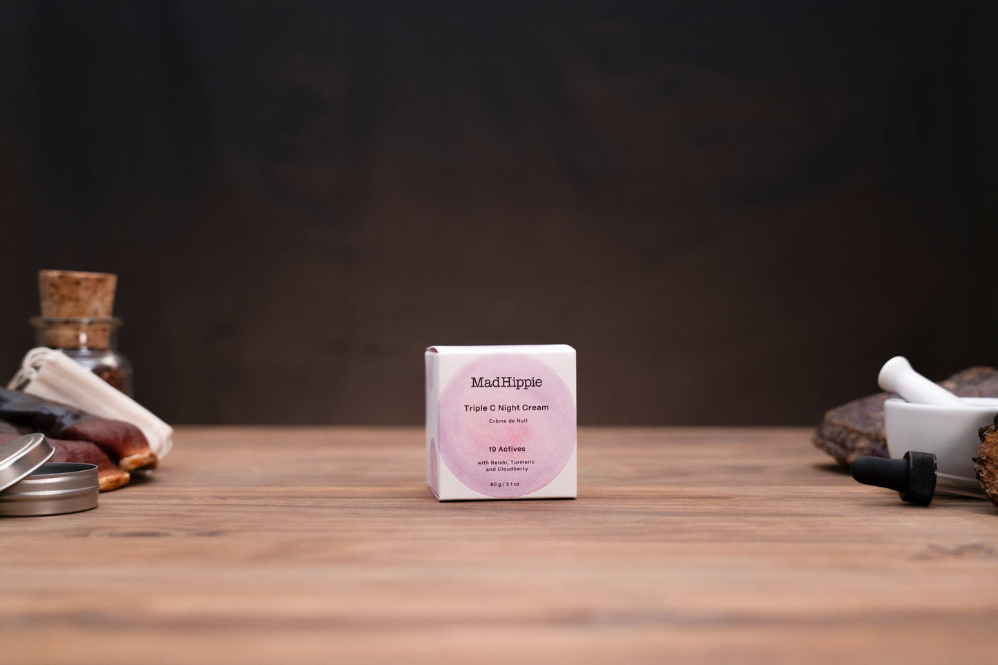 A small white square box of MatRipple Fragrance Oil Concentrate centered on a wooden tabletop, with a dark blurred background and decorative objects partially visible at the sides.