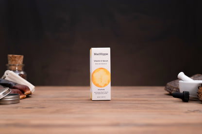 A small white box of MacHippie Vitamin E Serum stands centered on a wooden tabletop against a dark brown background, with blurred cosmetic props on both sides.