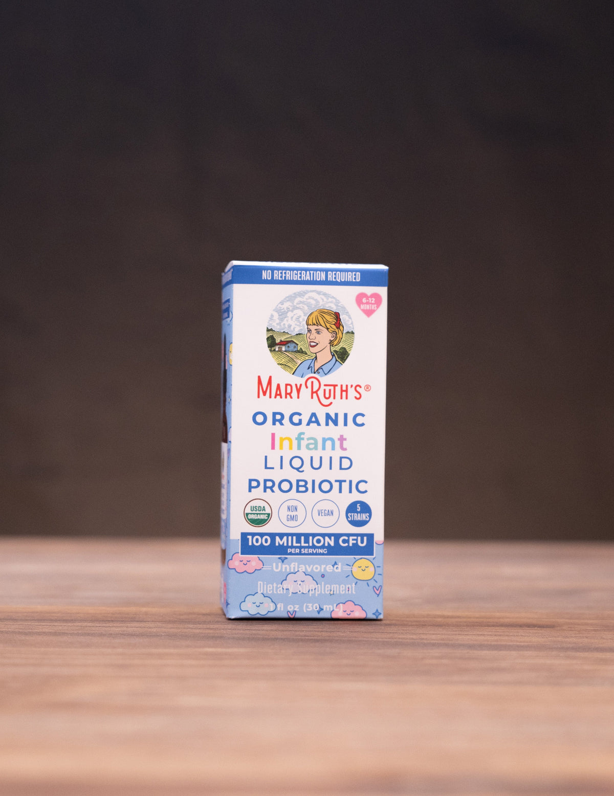 A box of Mary's Organic Infant Liquid Probiotic stands centered on a wooden surface against a dark backdrop, with small styled props visible at the left and right edges.