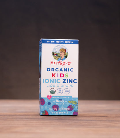 A box of Mary's Organic Kids Ionic Zinc Liquid Drops stands centered on a wooden tabletop against a dark backdrop. The package is white with blue and purple text and graphics, and the brand Mary's is clearly visible.