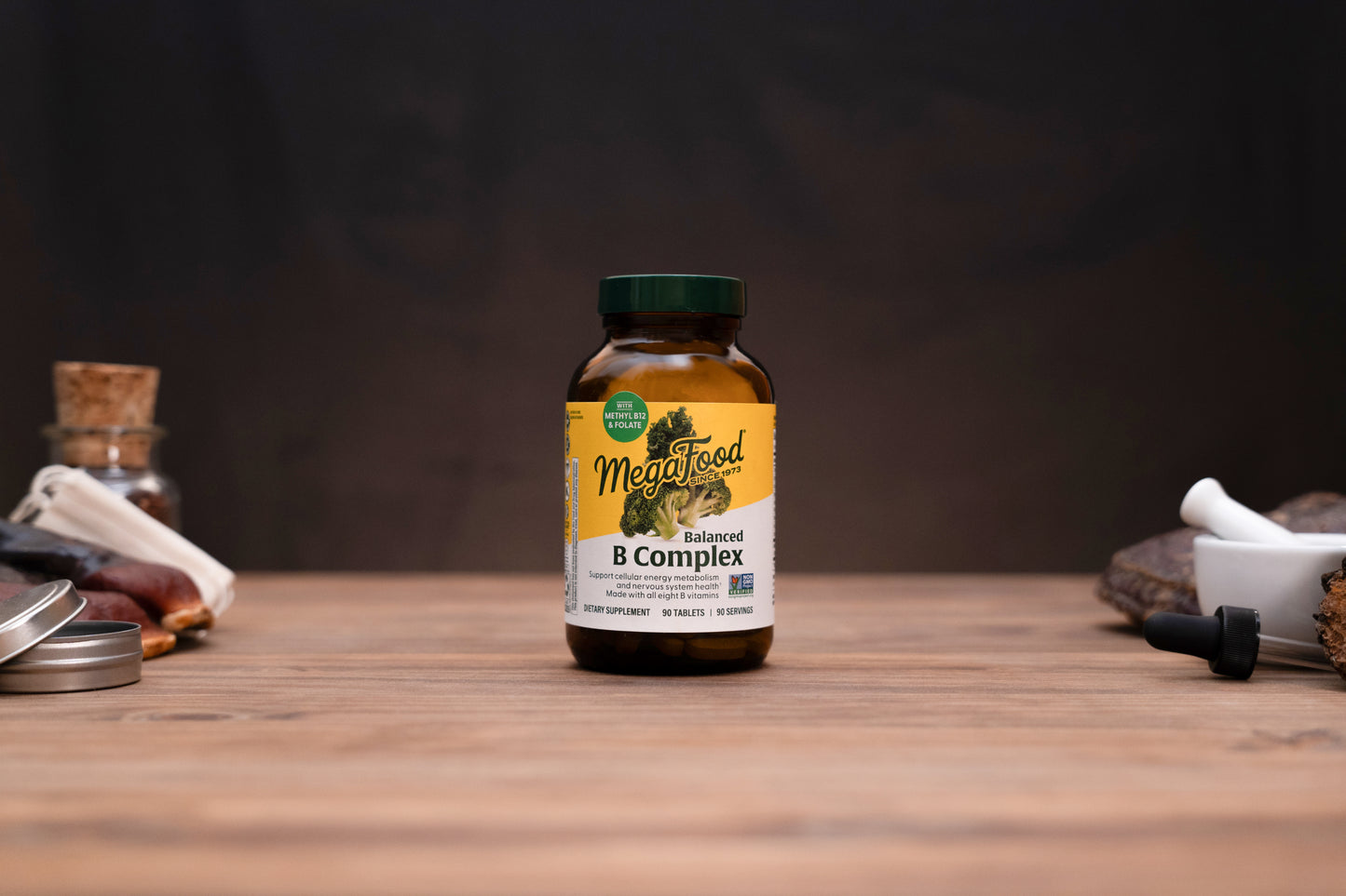A bottle of MegaFood Sustained B Complex dietary supplement standing on a wooden tabletop, with blurred herbal and kitchen props on both sides against a dark background.