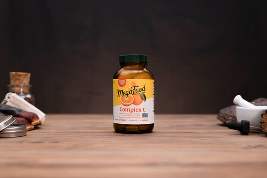 A bottle of MegaFood Complex C dietary supplement centered on a wooden tabletop, with blurred herbal props on both sides against a dark background.