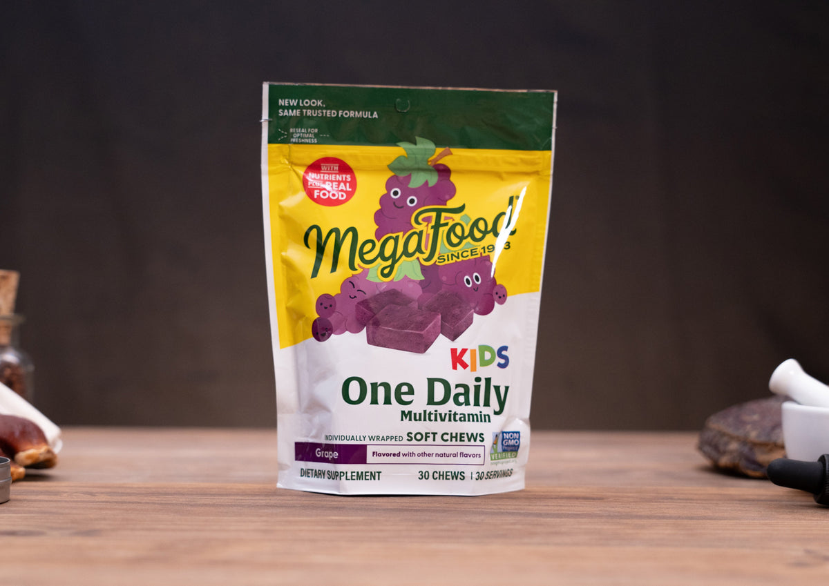 A stand-up pouch of MegaFood Kids One Daily Multivitamin soft chews, grape flavor, displayed on a wooden surface with blurred rustic props in the background.