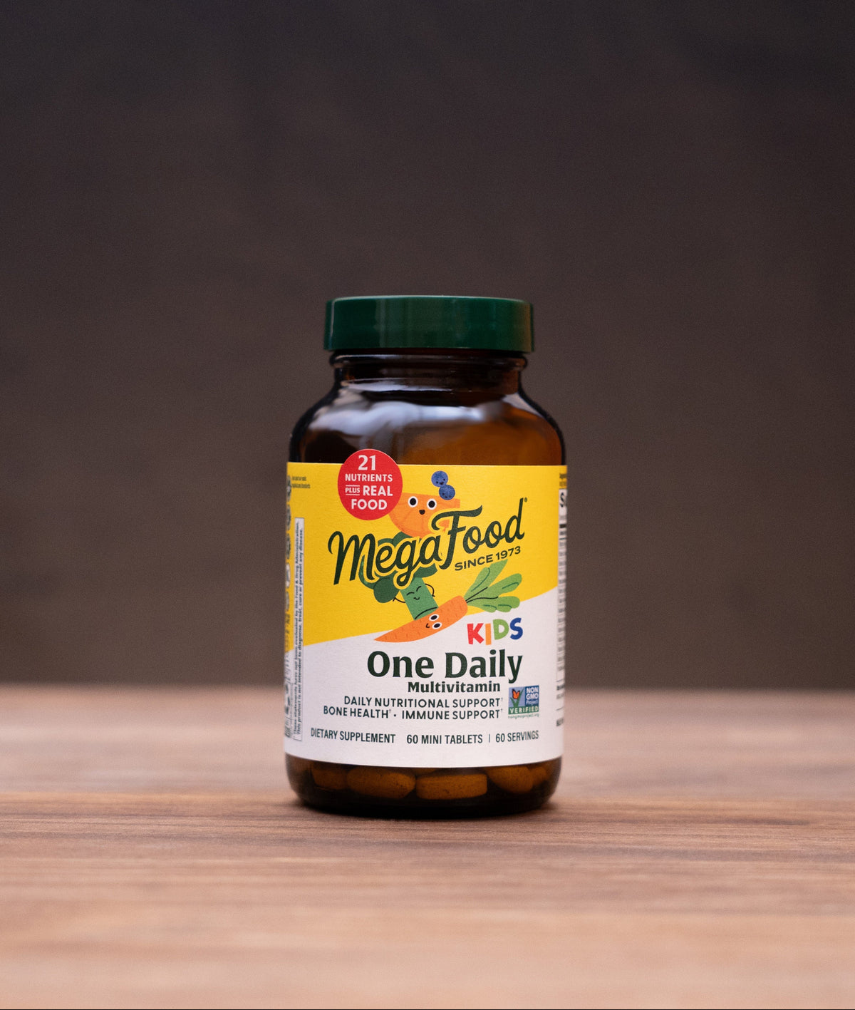 A bottle of MegaFood One Daily supplement centered on a wooden tabletop, with blurred herbal ingredients and a mortar and pestle in the background.