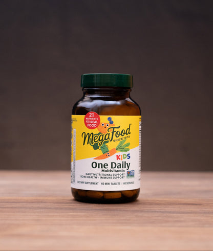 A bottle of MegaFood One Daily supplement centered on a wooden tabletop, with blurred herbal ingredients and a mortar and pestle in the background.