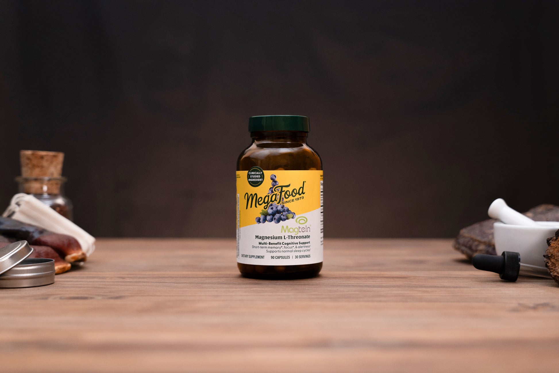A bottle of MegaFood Magnesium Citrate supplement standing on a wooden surface against a dark background, with small blurred props on both sides.