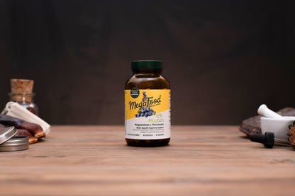 A bottle of MegaFood Magnesium Citrate supplement standing on a wooden surface against a dark background, with small blurred props on both sides.