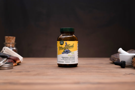 A bottle of MegaFood Magnesium Citrate supplement standing on a wooden surface against a dark background, with small blurred props on both sides.