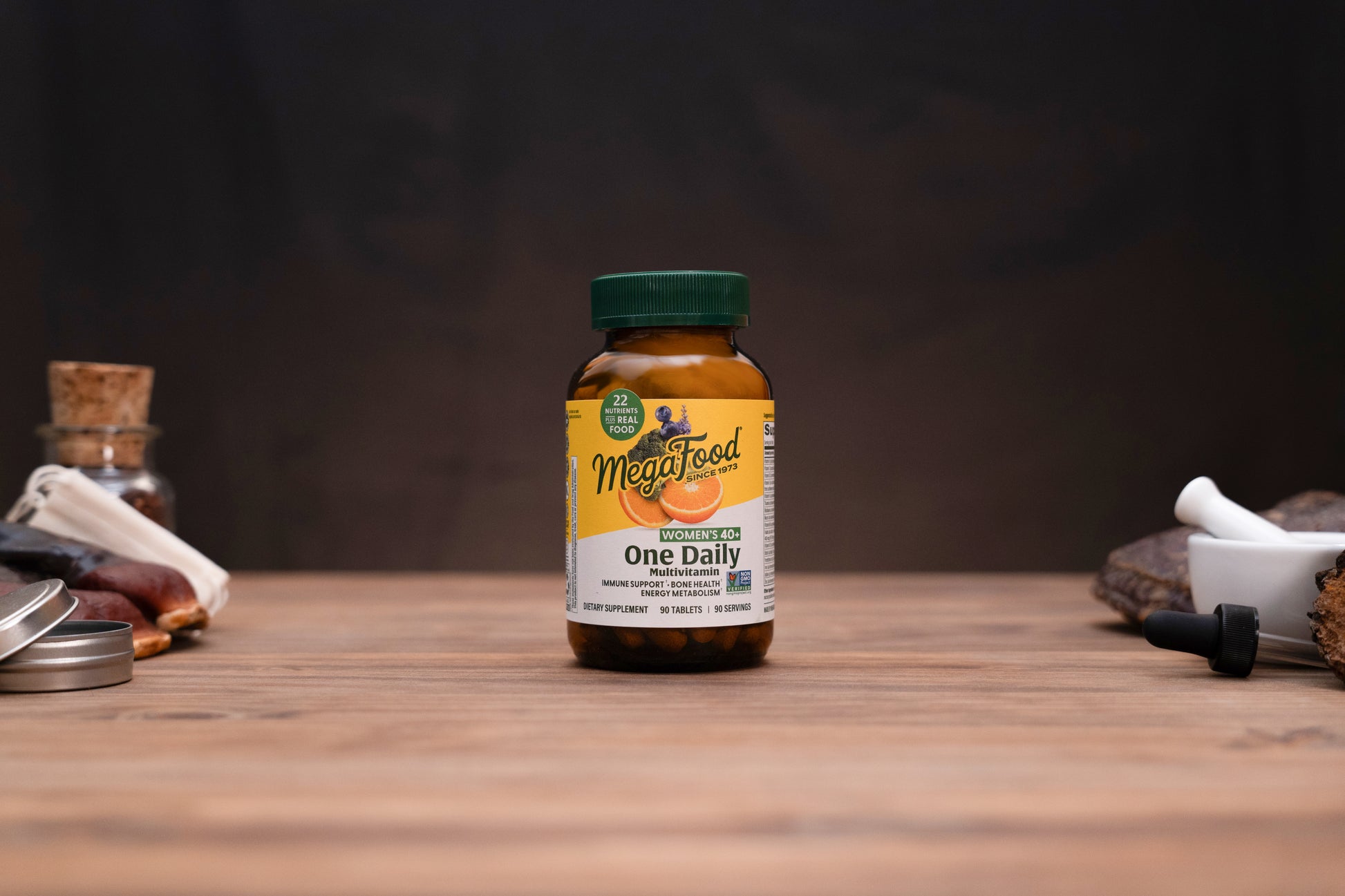 A bottle of MegaFood One Daily dietary supplement centered on a wooden tabletop, with blurred herbal props and a dark background.