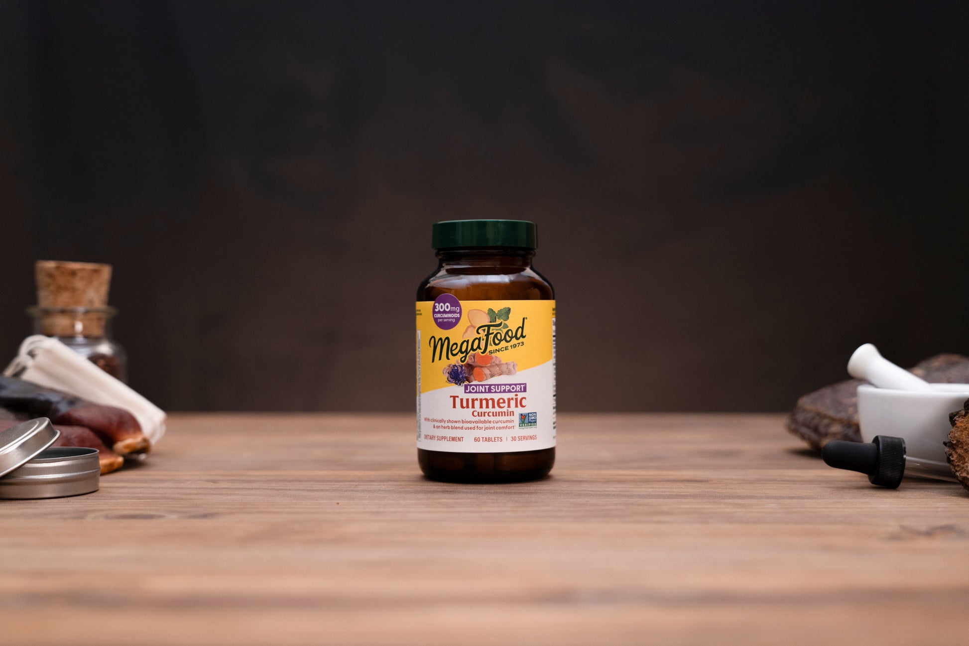 A bottle of MegaFeed Turmeric dietary supplement centered on a wooden tabletop, with blurred herbal props and a dark background.