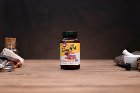 A bottle of MegaFeed Turmeric dietary supplement centered on a wooden tabletop, with blurred herbal props and a dark background.