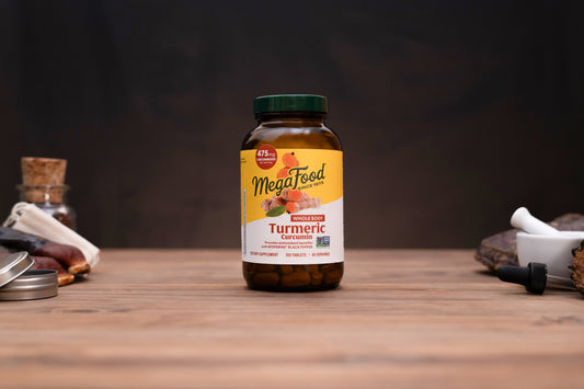 A bottle of MegaFood Turmeric Curcumin dietary supplement standing on a wooden surface against a dark background, with small blurred herbal props on both sides.