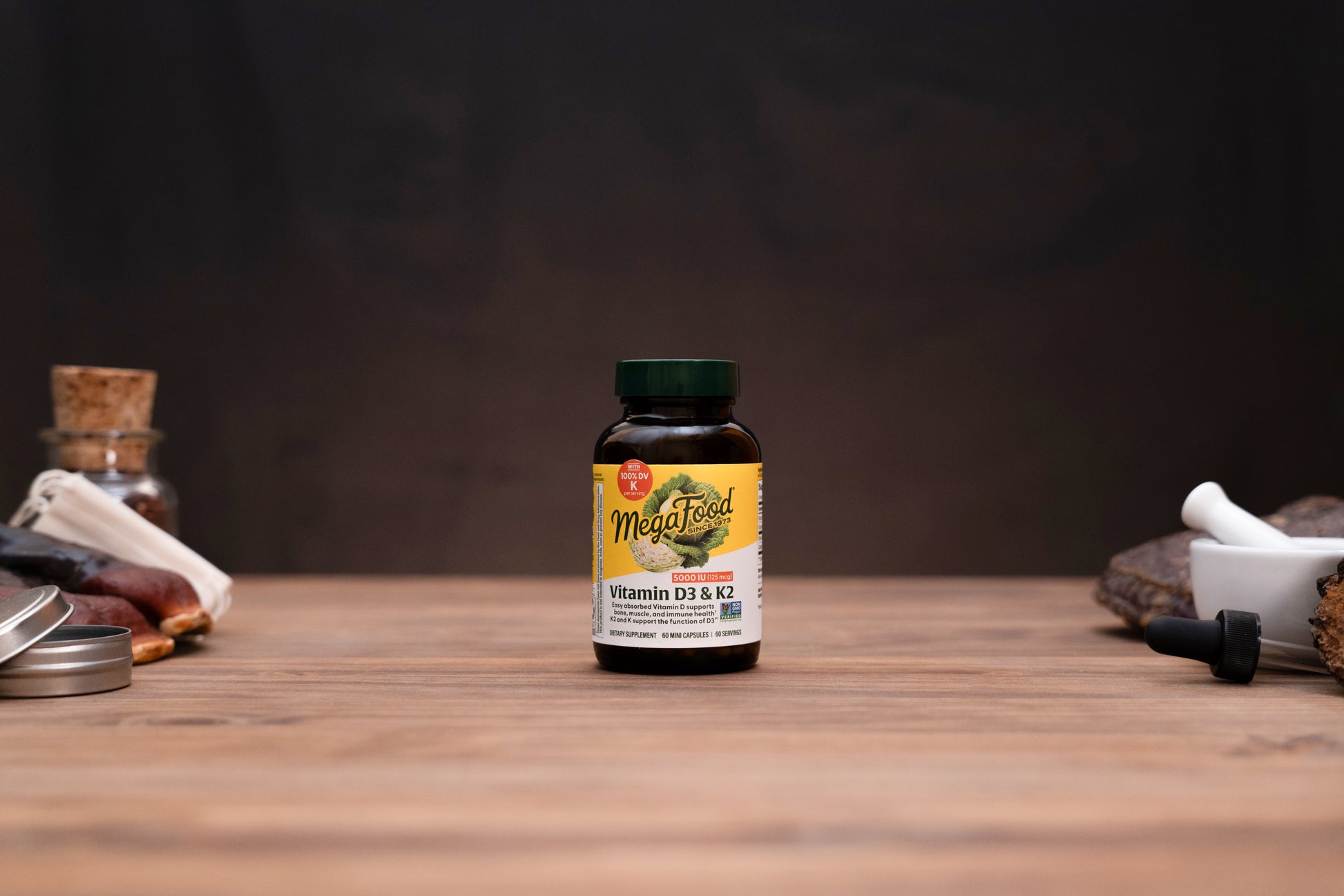 A bottle of MegaFood Vitamin D3 & K2 dietary supplement standing on a wooden tabletop, with blurred apothecary-style props on both sides against a dark background.