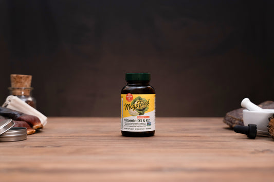 A bottle of MegaFood Vitamin D3 & K2 dietary supplement standing on a wooden tabletop, with blurred apothecary-style props on both sides against a dark background.