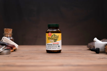 A bottle of MegaFeed Vitamin D3 & K2 dietary supplement standing on a wooden surface against a dark background, with blurred herbal props on either side.
