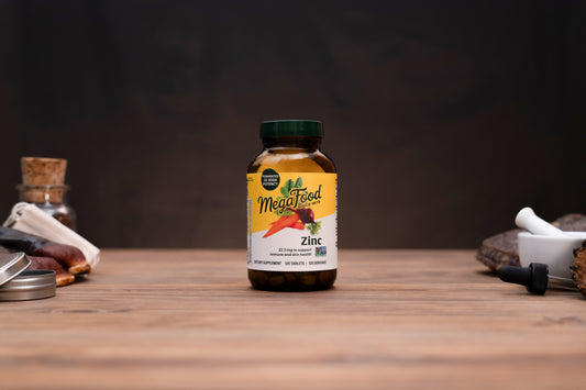 A bottle of MegaFood Zinc dietary supplement standing on a wooden tabletop, with blurred herbal props on both sides against a dark background.