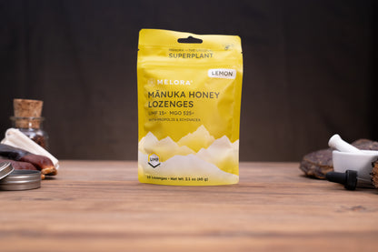 A yellow stand-up pouch of Melora Mánuka Honey Lozenges, Lemon flavor, standing on a wooden tabletop with rustic props blurred in the background.