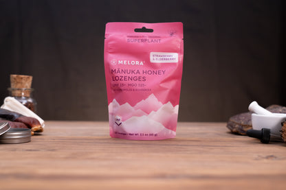 A pink stand-up pouch of Melora Mânuka Honey Lozenges on a wooden tabletop with blurred natural props in the background.