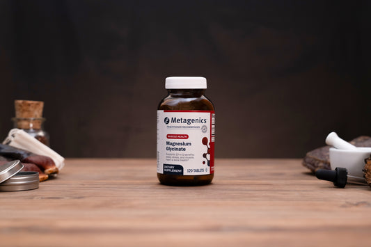 A bottle of Metagenics Magnesium Oxinate dietary supplement standing on a wooden tabletop, with blurred herbal props in the background.