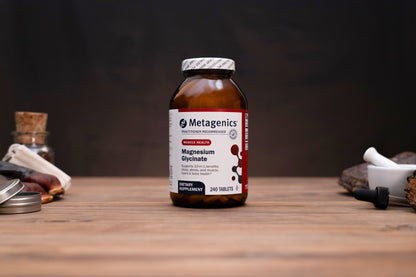 A bottle of Metagenics Magnesium Glycinate dietary supplement, set on a wooden tabletop with a dark, moody background and subtle herbal props.