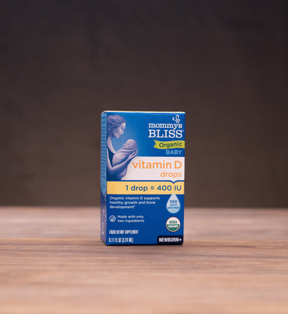 A box of Morinda Bliss Vitamin D drops stands centered on a wooden tabletop against a dark backdrop, with blurred rustic props on both sides.