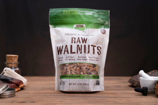 A 12 oz bag of NOW Foods Raw Walnuts, organic and unprocessed, standing on a wooden surface with a dark background and small kitchen props on either side.