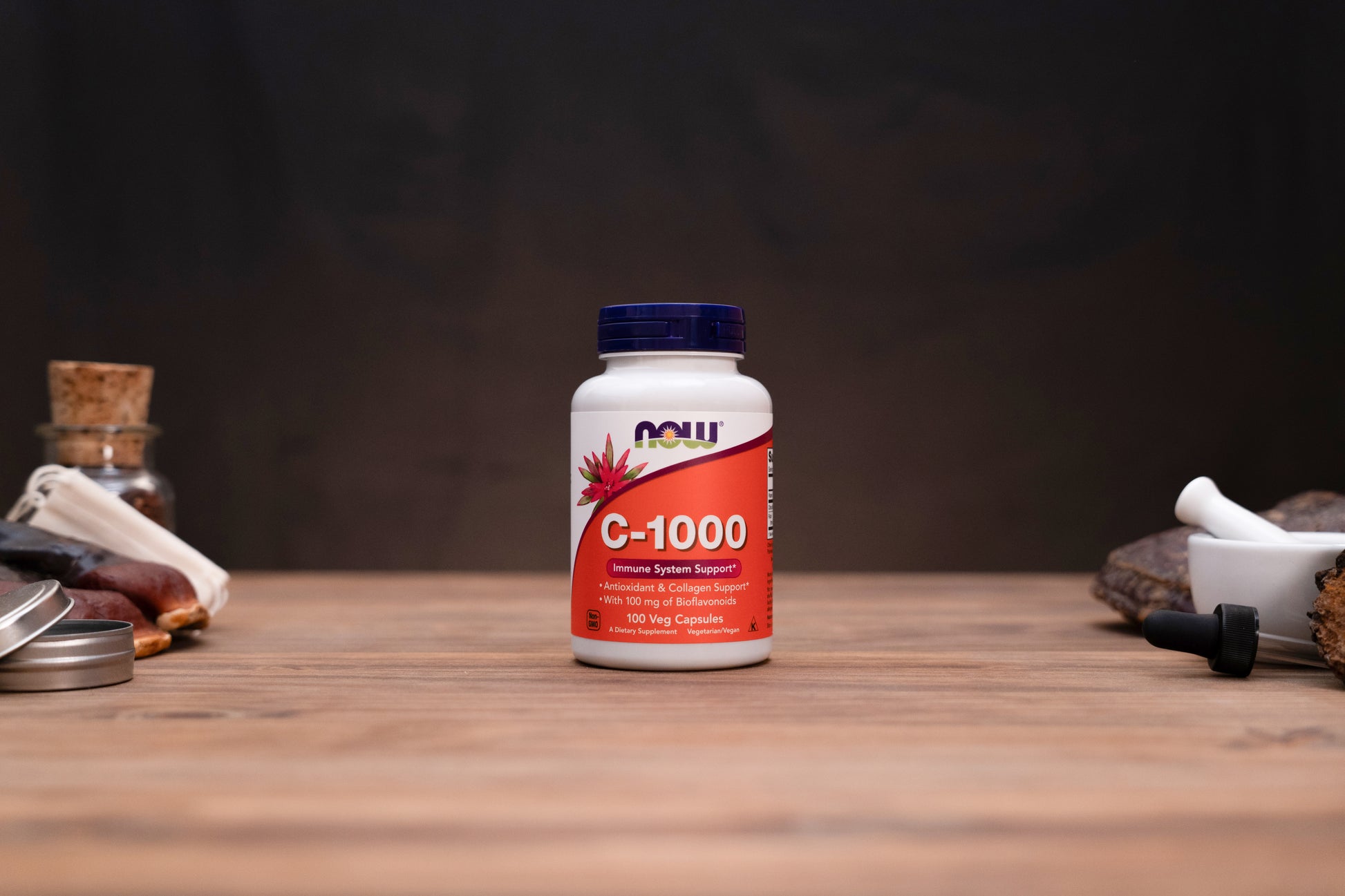 A bottle of NOW C-1000 dietary supplement capsules standing on a wooden tabletop against a dark background, with small apothecary-style props visible on both sides.