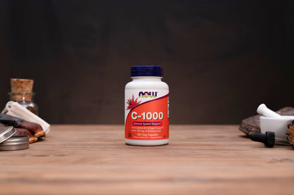 A bottle of NOW C-1000 dietary supplement capsules standing on a wooden tabletop against a dark background, with small apothecary-style props visible on both sides.