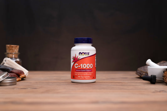 A bottle of NOW C-1000 dietary supplement capsules standing on a wooden tabletop against a dark background, with small apothecary-style props visible on both sides.