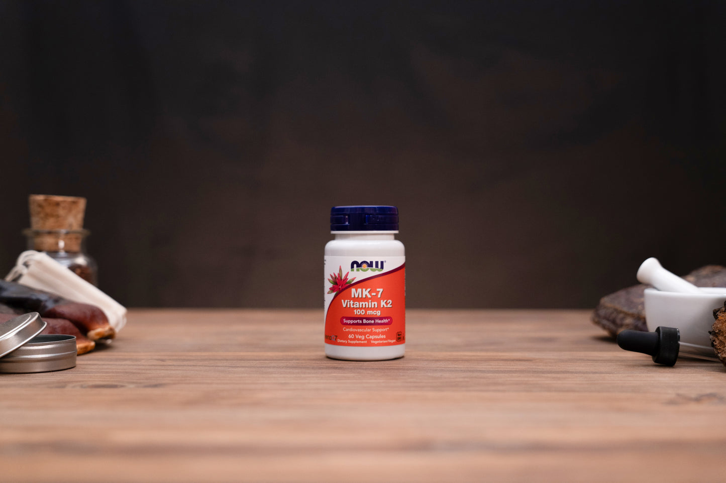 A bottle of NOW Vitamin K2 supplement centered on a wooden tabletop, with blurred herbal props on both sides against a dark background.