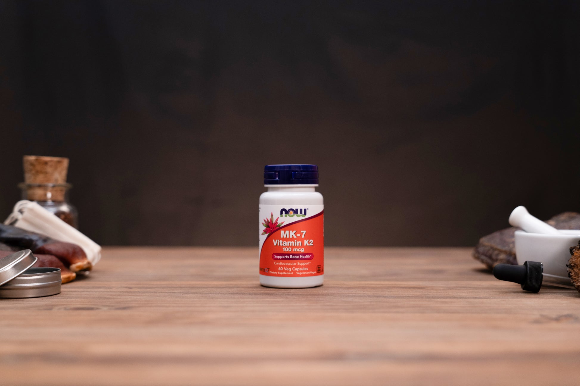 A bottle of NOW Vitamin K2 supplement centered on a wooden tabletop, with blurred herbal props on both sides against a dark background.