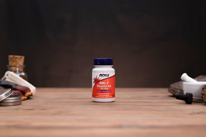 A bottle of NOW Vitamin K2 supplement centered on a wooden tabletop, with blurred herbal props on both sides against a dark background.