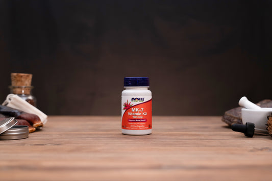 A bottle of NOW Vitamin K2 supplement centered on a wooden tabletop, with blurred herbal props on both sides against a dark background.