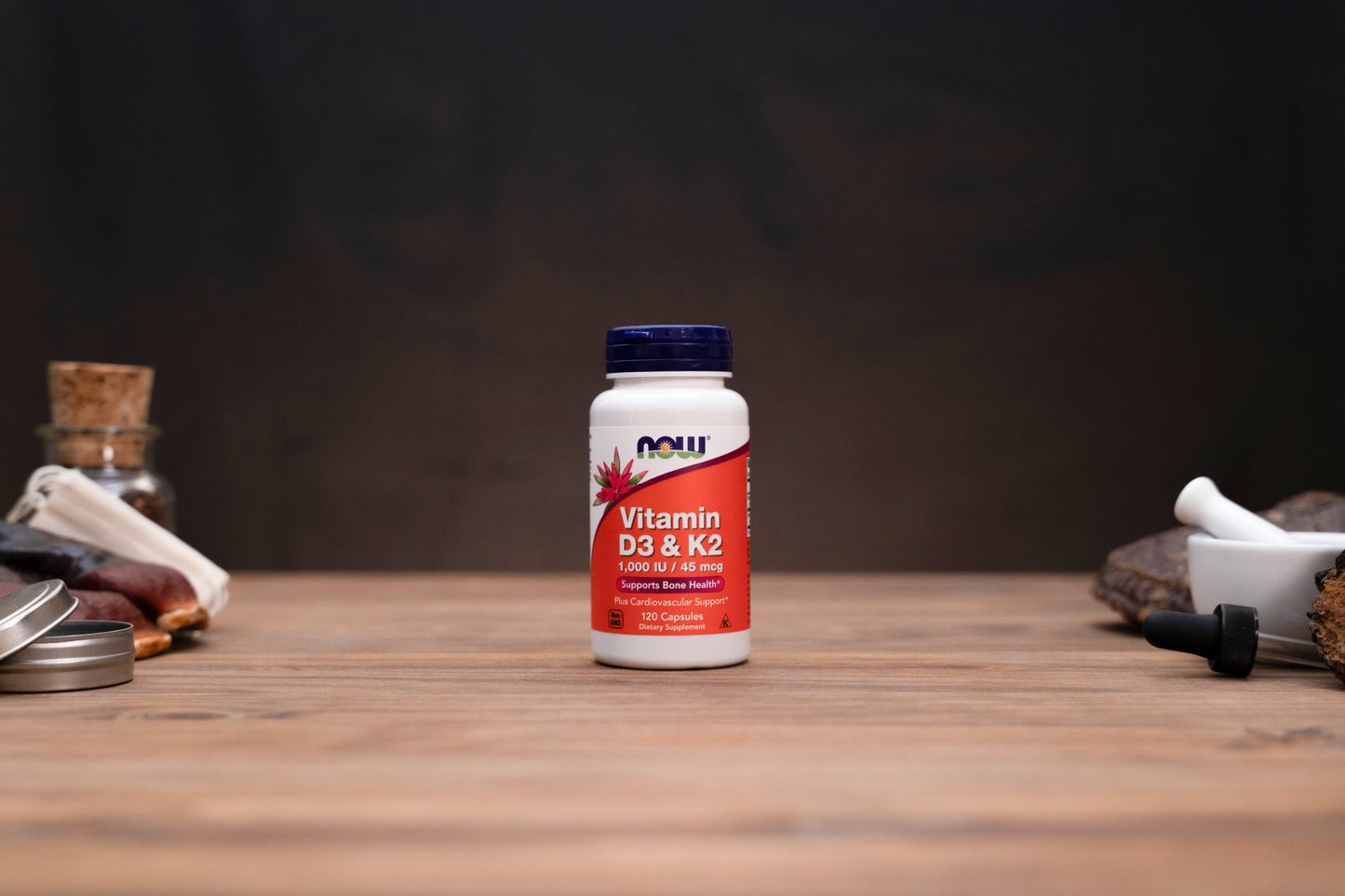 A bottle of NOW Vitamin D3 & K2 dietary supplement standing on a wooden tabletop, with blurred rustic props on both sides against a dark background.