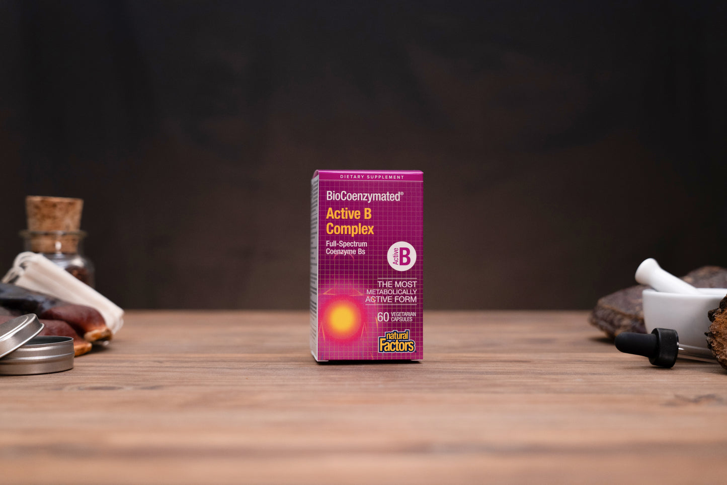 A box of BioConcentrated Active B Complex dietary supplement by Super Factory, standing on a wooden table with a dark, moody background and small herbal props on either side.