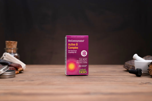A box of BioConcentrated Active B Complex dietary supplement by Super Factory, standing on a wooden table with a dark, moody background and small herbal props on either side.