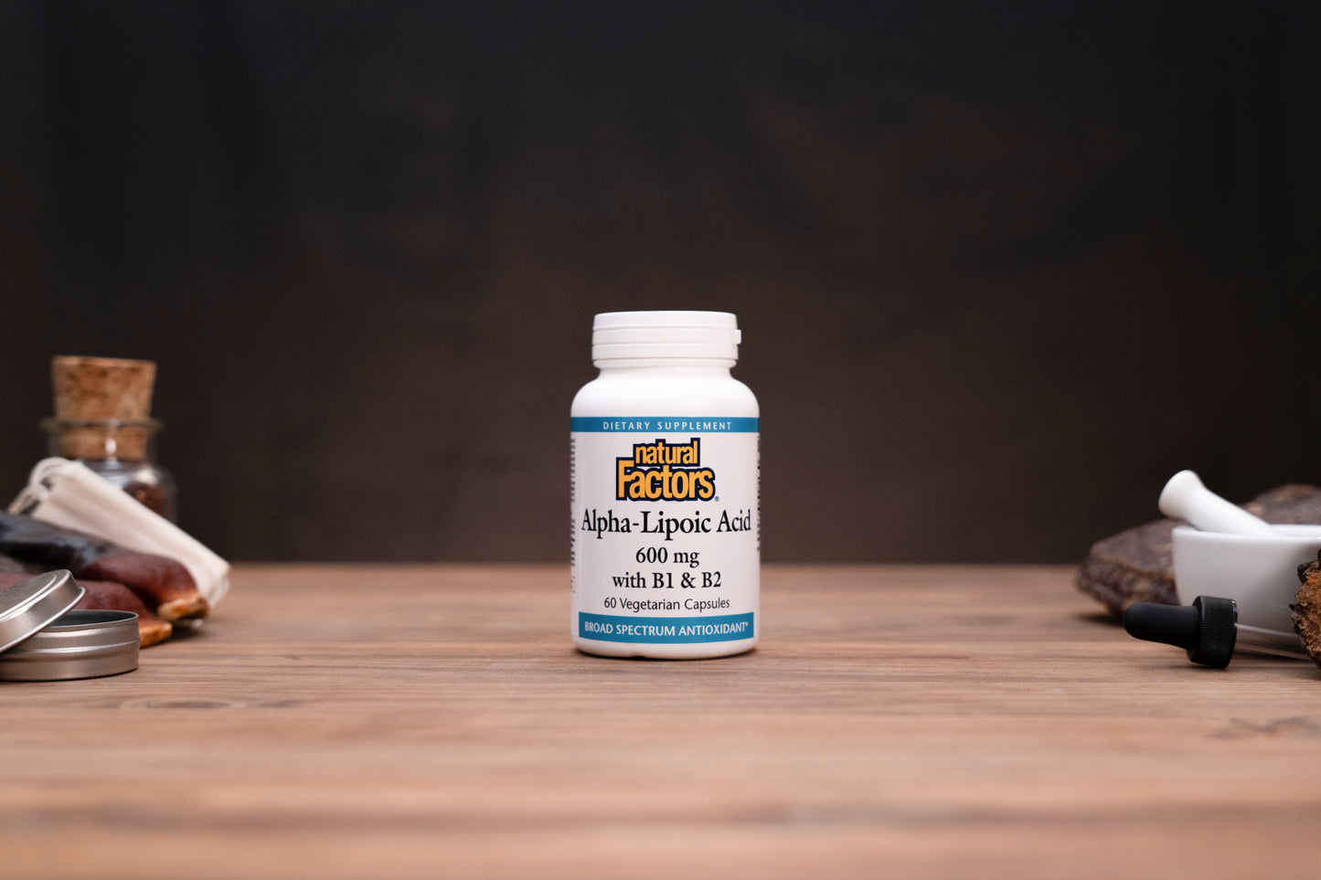 A white bottle of Natural Factors Alpha-Lipoic Acid 600 mg capsules stands on a wooden surface against a dark background, styled with small herbal props on both sides.